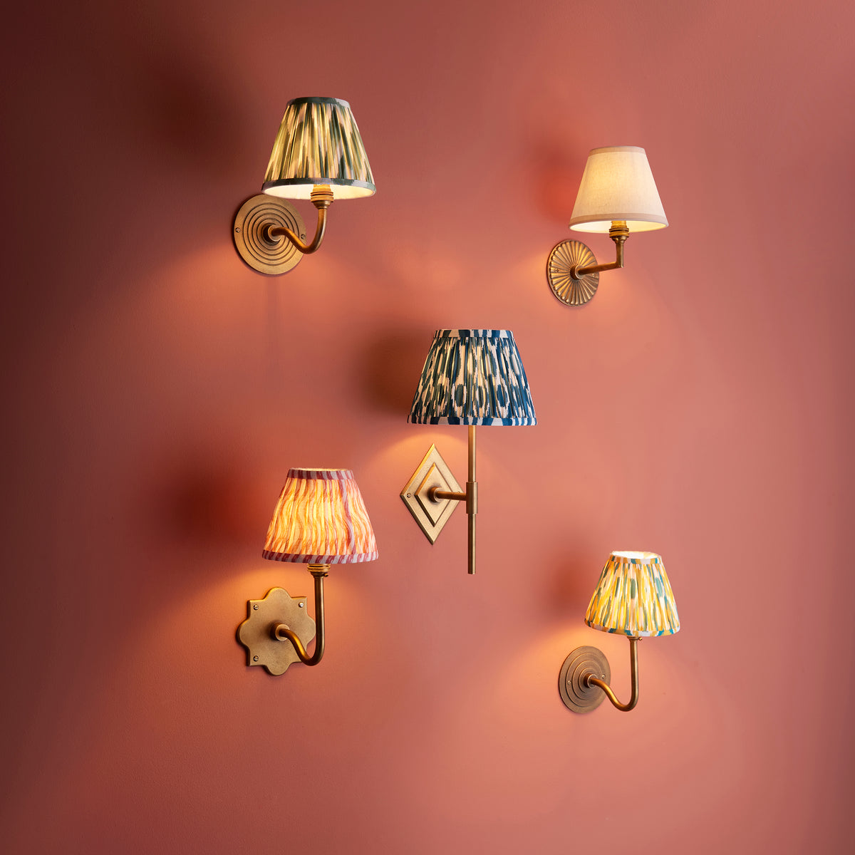 Five brass wall lights with assorted pleated ikat and plain shades grouped on a terracotta wall