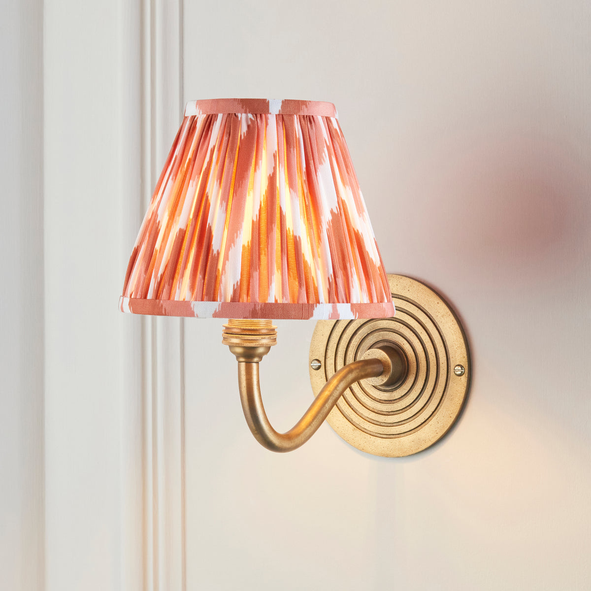 Ripple Curve brass wall light lit on panelled wall, pink-orange ikat pleated shade glowing
