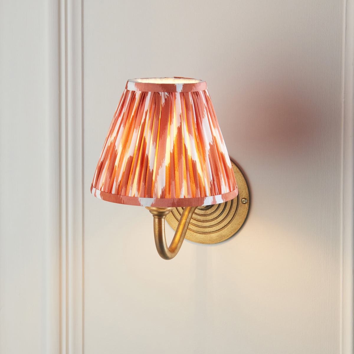 Ripple Curve brass wall light on a pale wall, pleated orange ikat shade seen from below