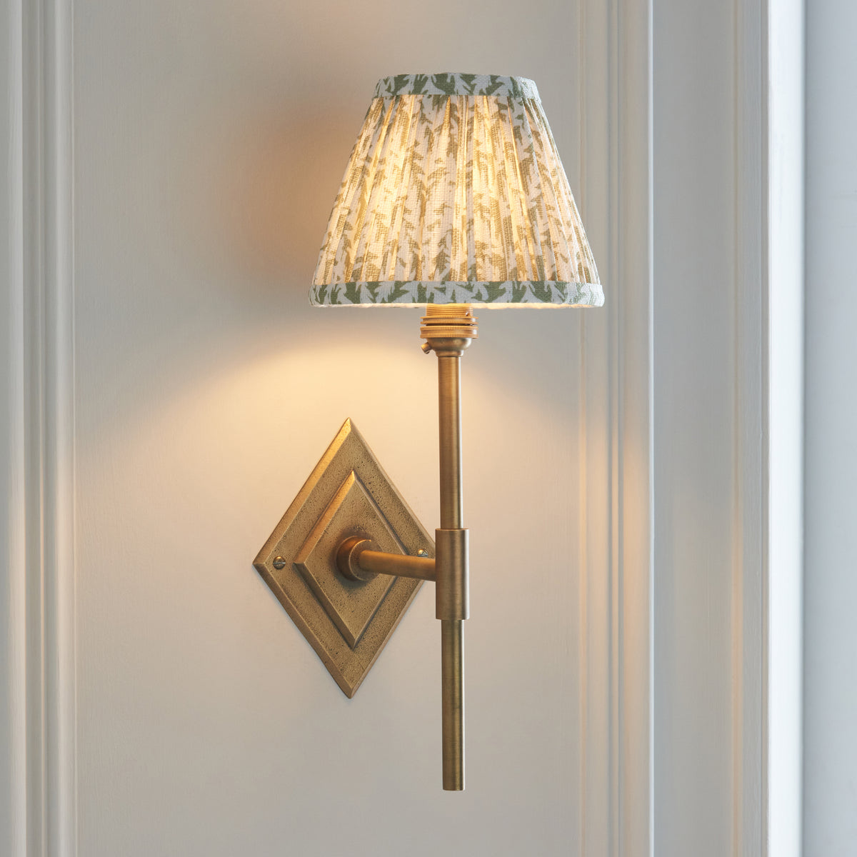 Diamond Tall antique brass wall light angled on pale panelling with warm leaf-print pleated shade