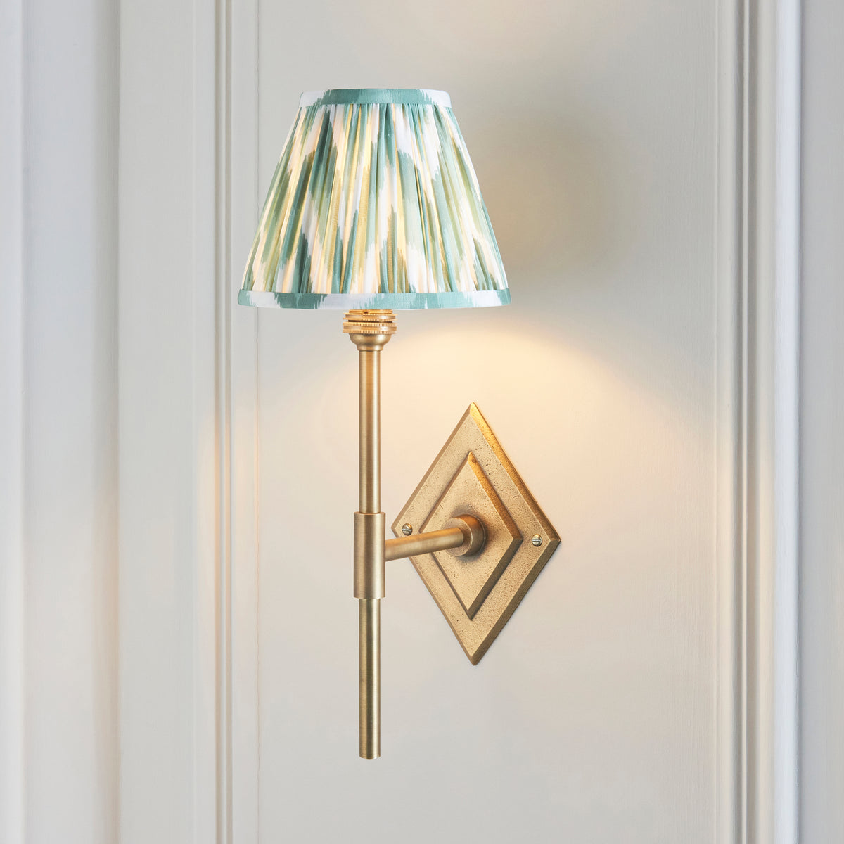 Diamond Tall brass wall light lit on panelled wall, green and cream zigzag ikat pleated shade