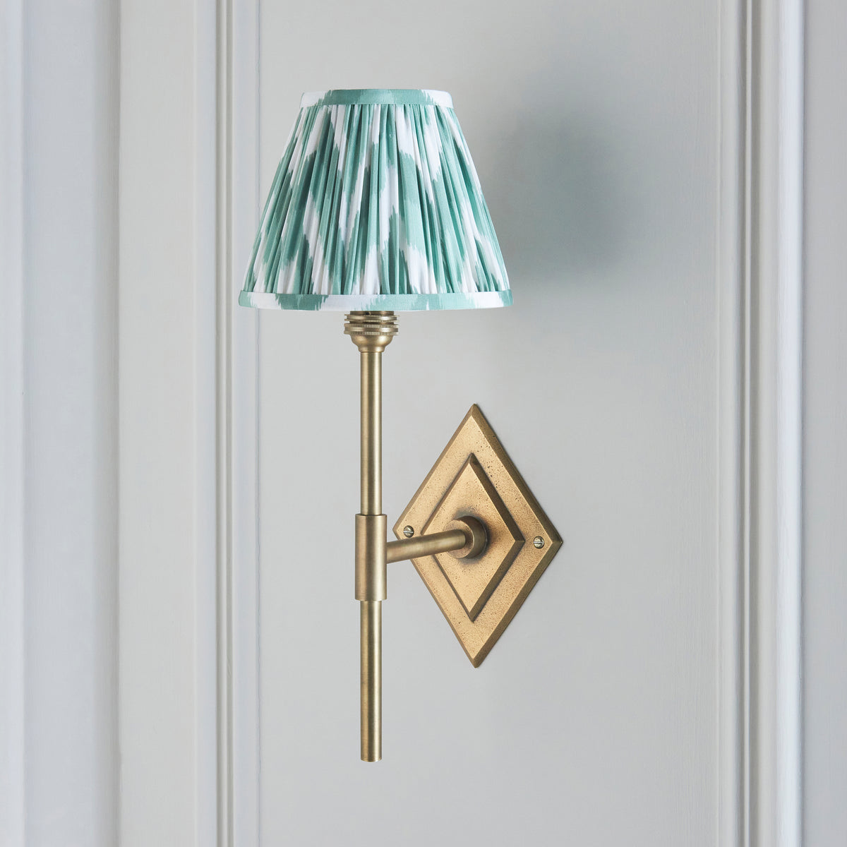 Diamond Tall brass wall light on a grey wall, softer green zigzag ikat pleated shade illuminated