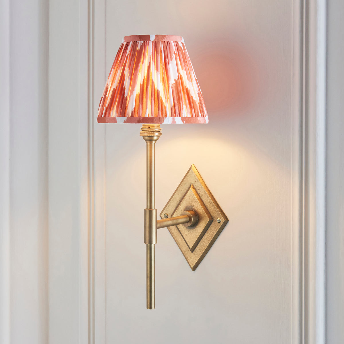 Diamond Tall brass wall light lit on panelled wall, pink-orange ikat pleated shade above a slim stem