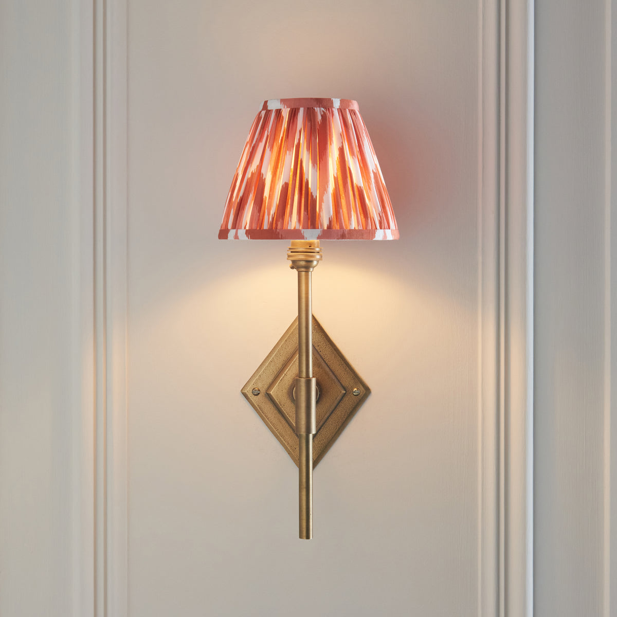 Diamond Tall brass wall light head-on against pale panelling with warm orange ikat pleated shade