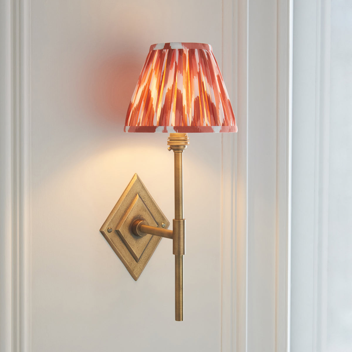 Diamond Tall antique brass wall light angled on off-white panelling, lit orange ikat shade