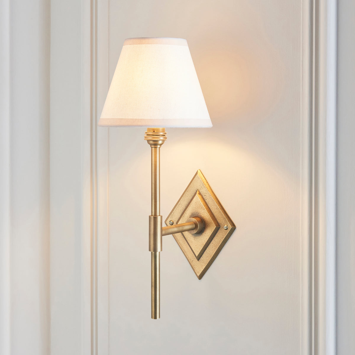 Diamond Tall brass wall light lit on panelled off-white wall, plain white fabric shade glowing