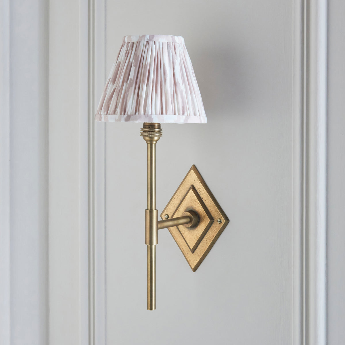 Diamond antique brass wall light lit against pale grey wall, softly pleated natural ikat shade