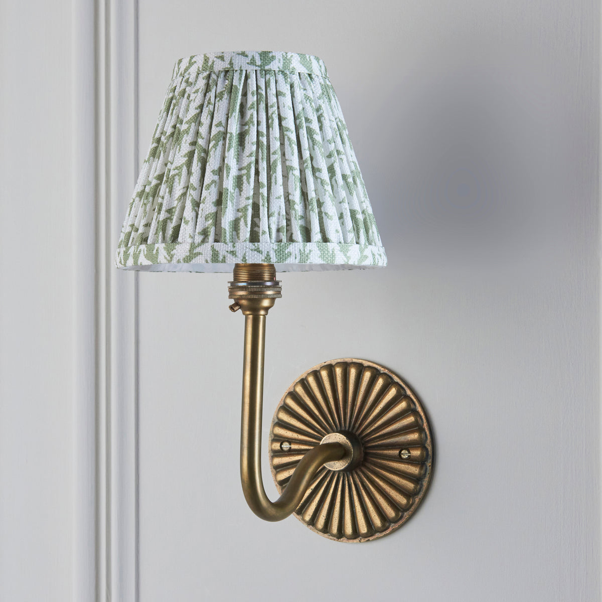 Dahlia Swan antique brass wall light against grey wall with green leaf-print pleated fabric shade