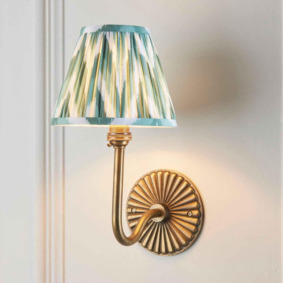 Dahlia Swan brass wall light lit on panelled wall, green ikat zigzag pleated shade glowing