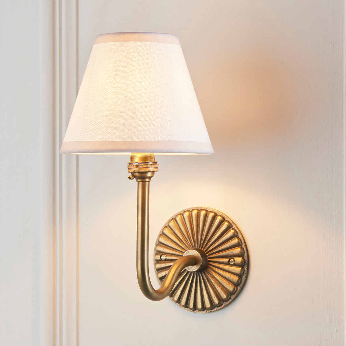 Dahlia Swan brass wall light lit on panelled wall, plain ivory linen tapered shade glowing