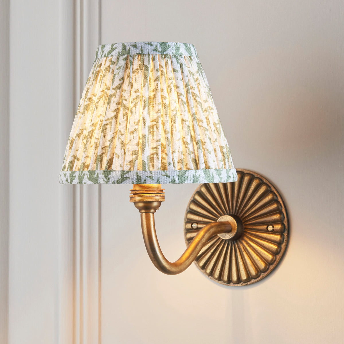 Dahlia Curve brass wall light lit on panelled wall, green leaf-print pleated shade glowing
