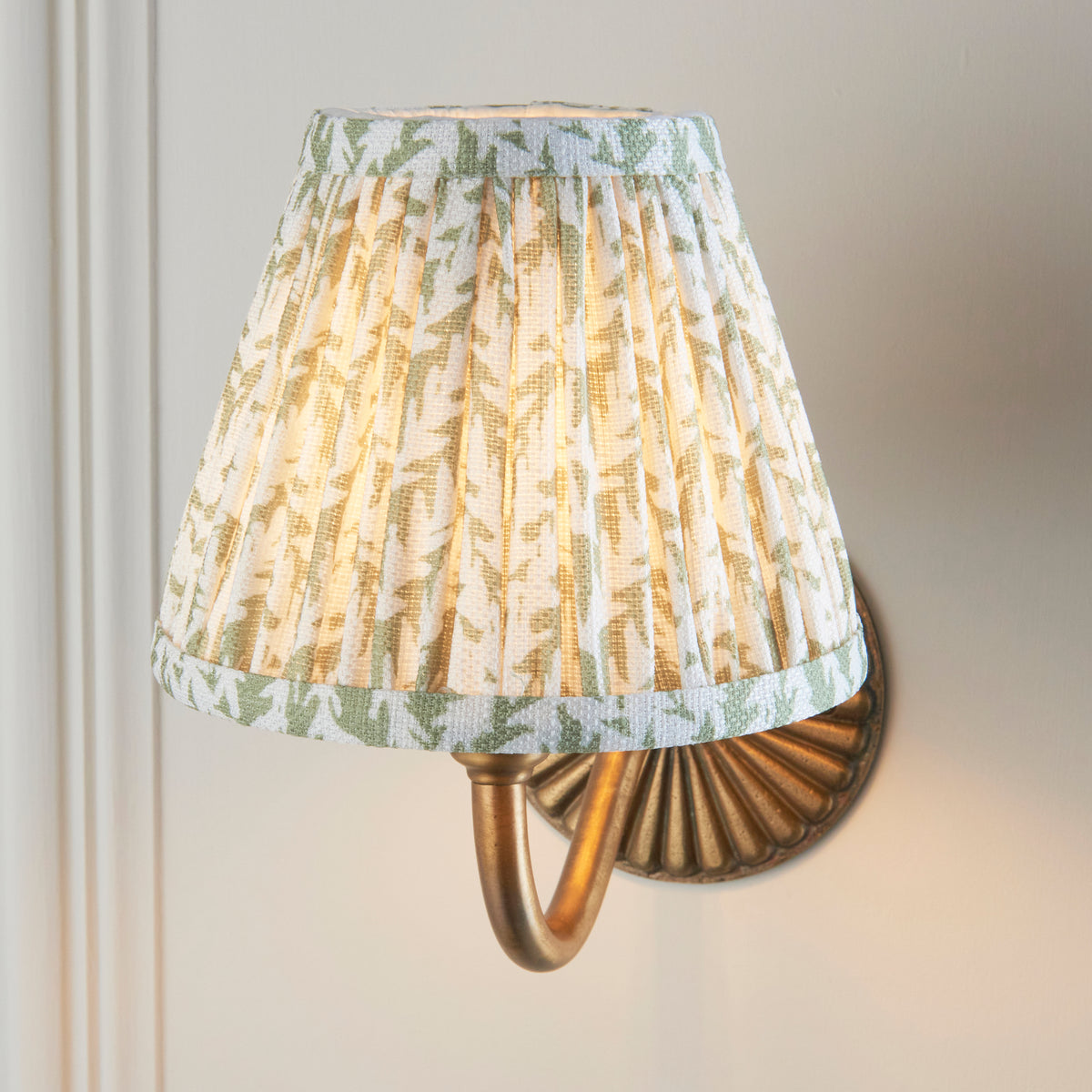 Green leaf-print pleated shade lit on the Dahlia Curve brass wall light against a cream wall