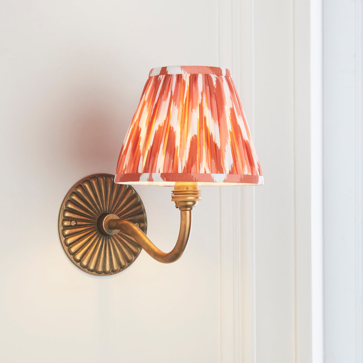 Angled view of the Dahlia Curve brass sconce casting warm light through its orange ikat pleated shade