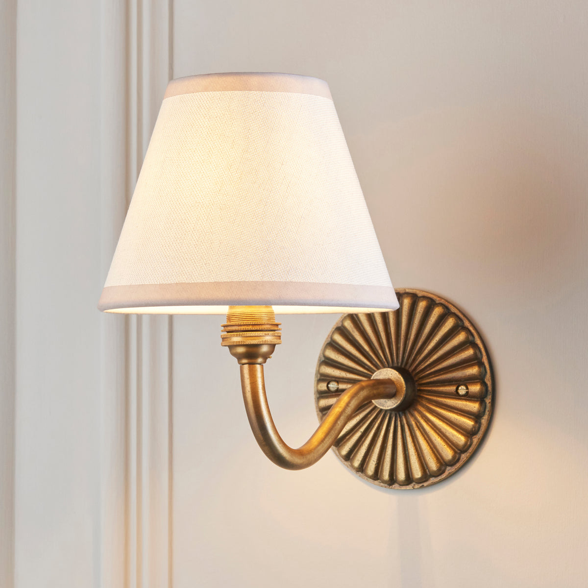Ivory fabric shade lit on the Dahlia Curve & Ivy brass wall light against pale wall panelling