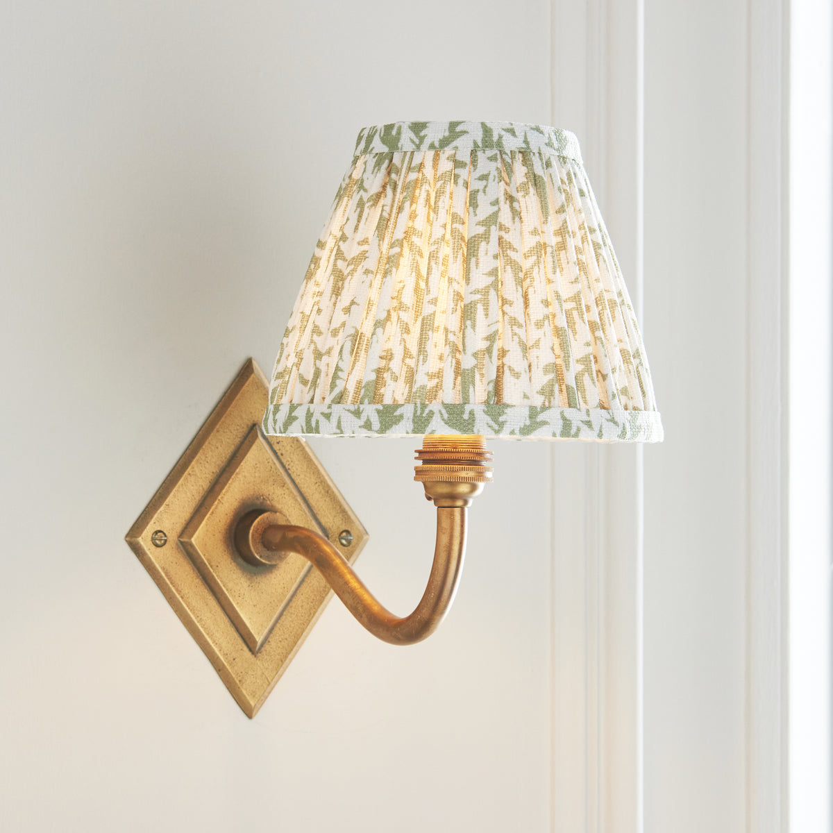 Angled view of the Diamond Curve brass wall light with softly lit green foliage patterned shade