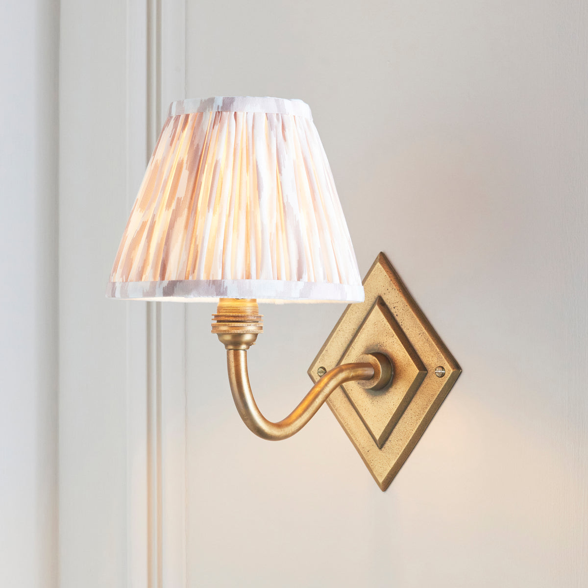 Natural ikat pleated shade glowing on the Diamond Curve brass wall light against pale panelling