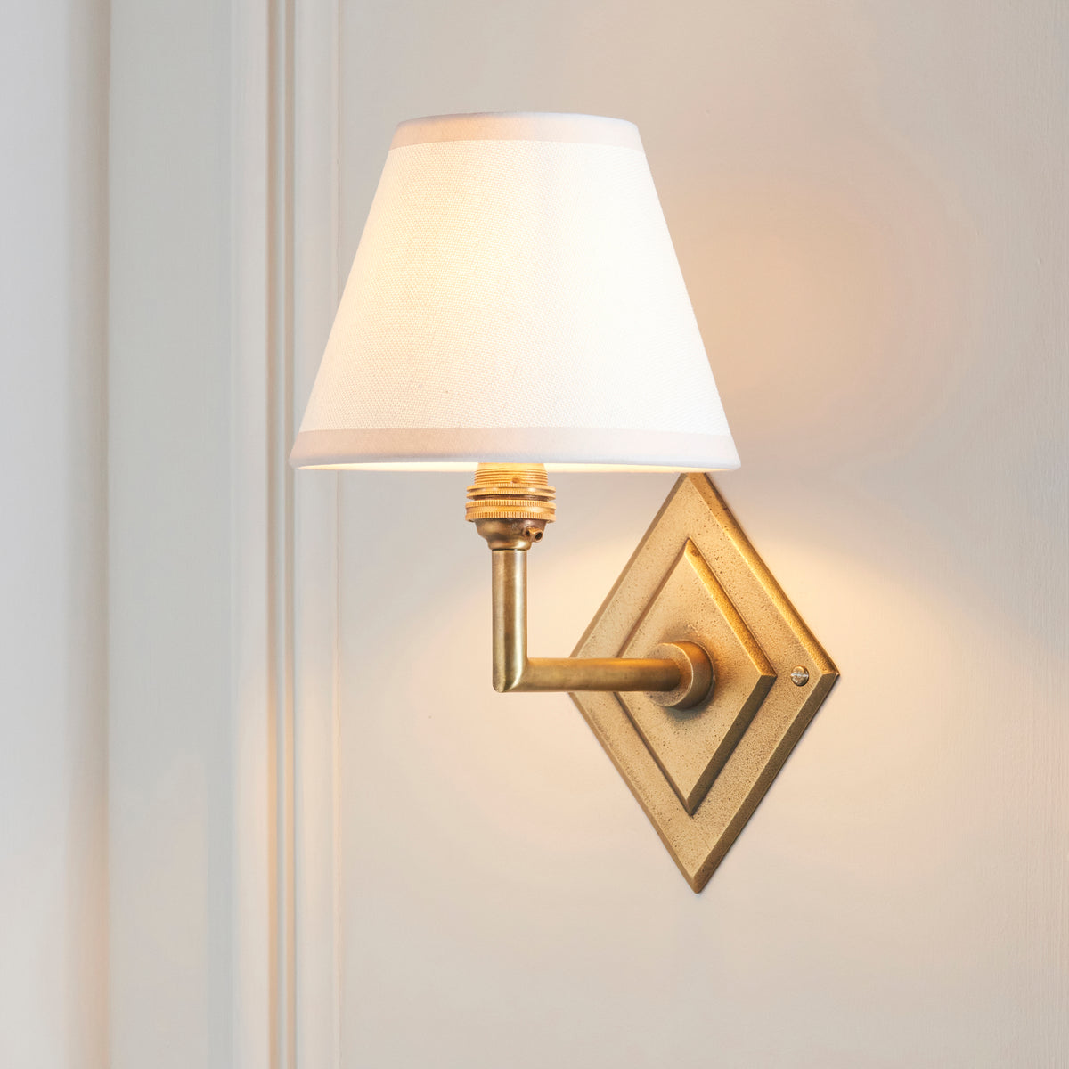 Diamond Angle & Ivy brass wall light with white shade, lit and angled on white panelling
