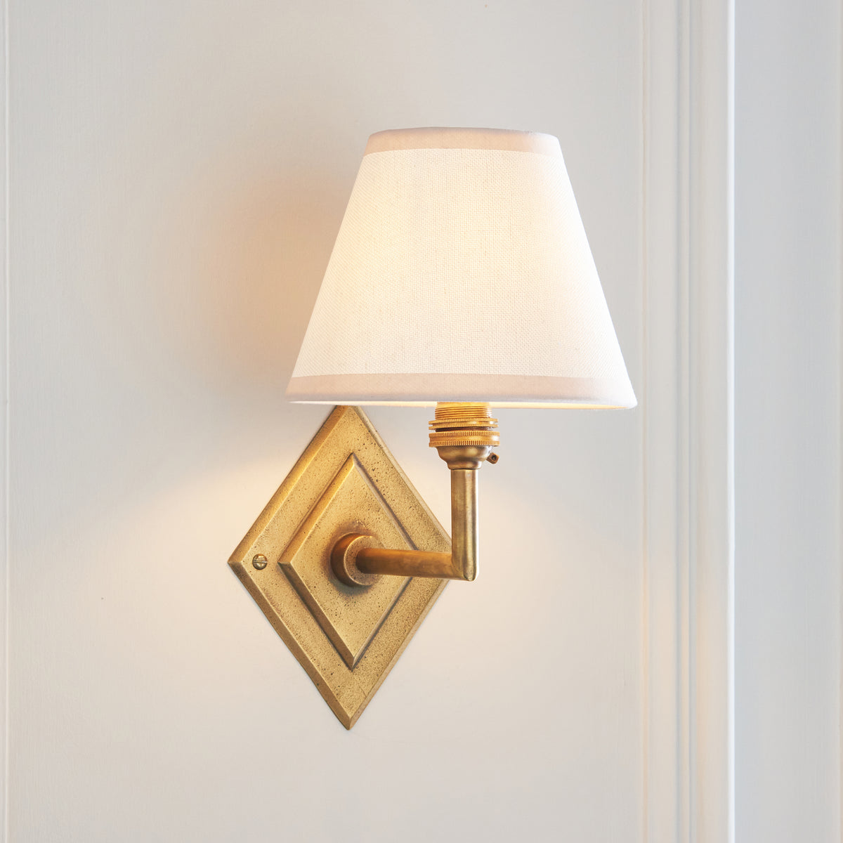 White-shaded Diamond Angle & Ivy brass wall light glowing on a panelled white wall
