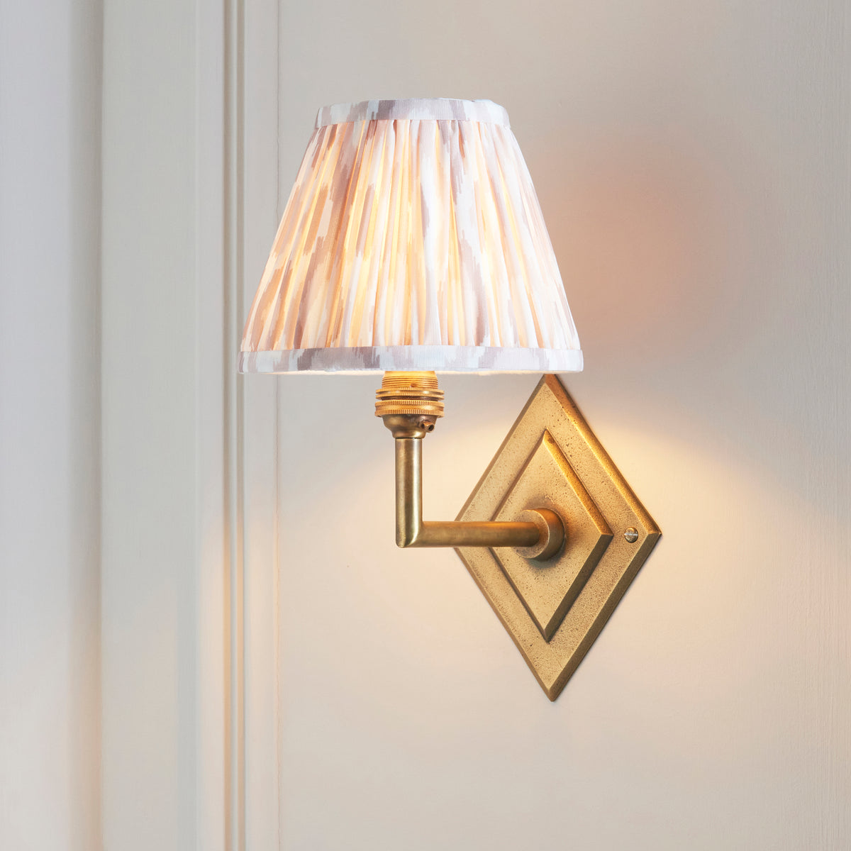 Diamond Angle brass wall light with natural ikat shade, lit at an angle on white panelling
