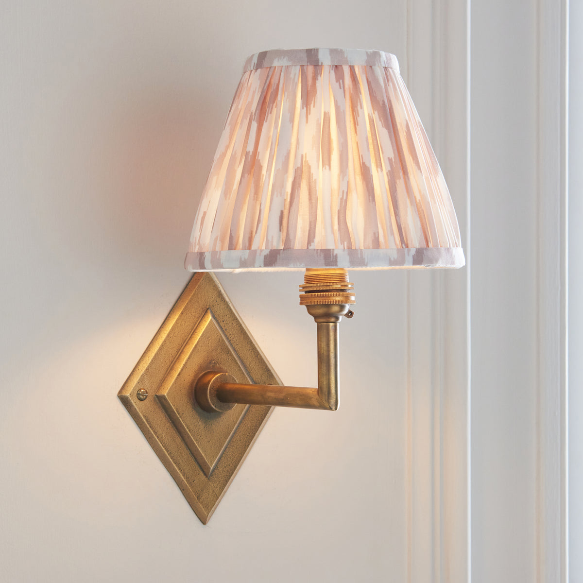 Natural ikat pleated shade on the Diamond Angle brass wall light, warmly lit on a white wall