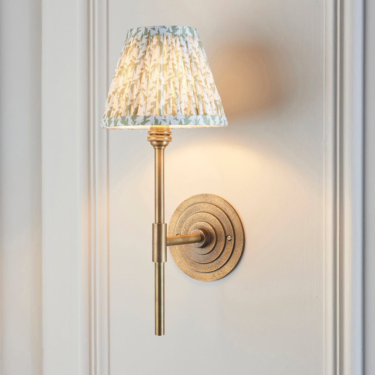 Halo brass wall light with green leaf pleated shade, lit at an angle on white panelling