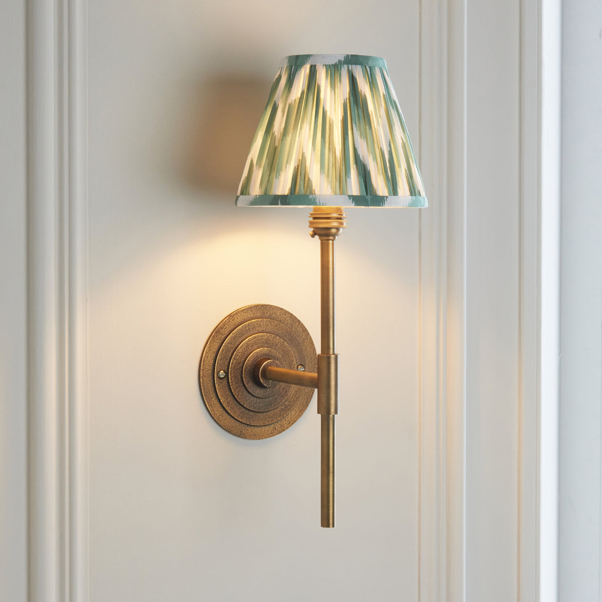 Halo Tall brass wall light glowing on a white panelled wall with green zigzag ikat shade