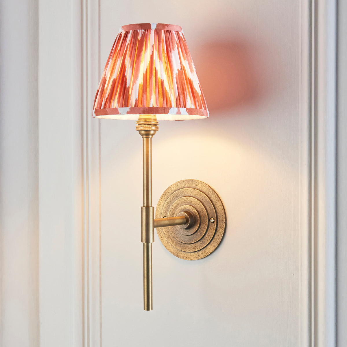 Halo brass wall light with orange ikat shade, lit at an angle on white panelling