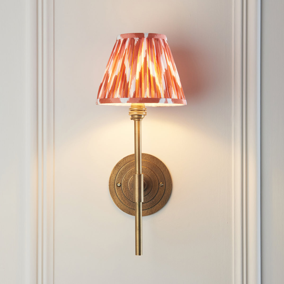 Lit Halo wall light head-on, brass stem and round plate beneath an orange ikat shade