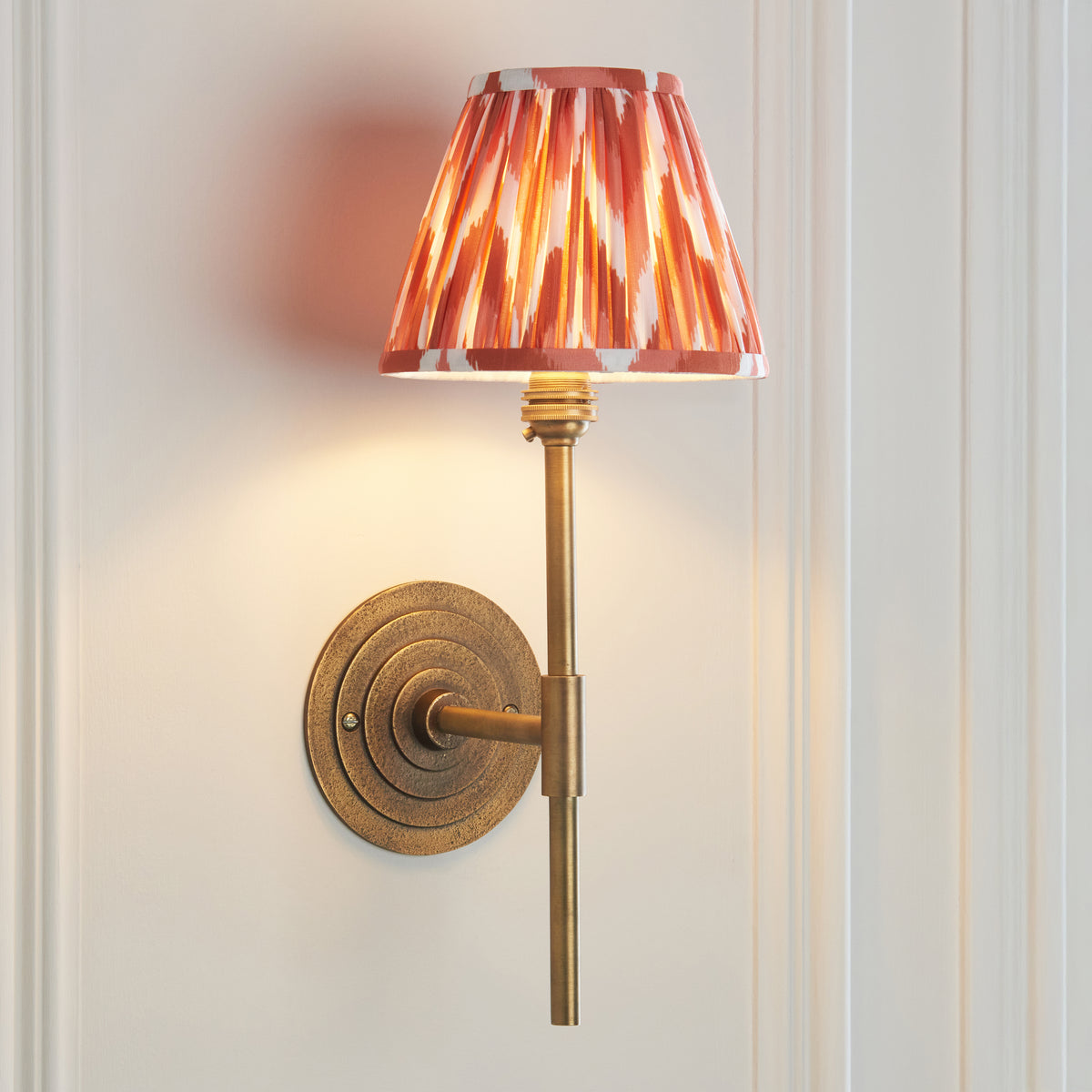 Halo brass wall light casting warm light on white panelling, orange zigzag ikat shade
