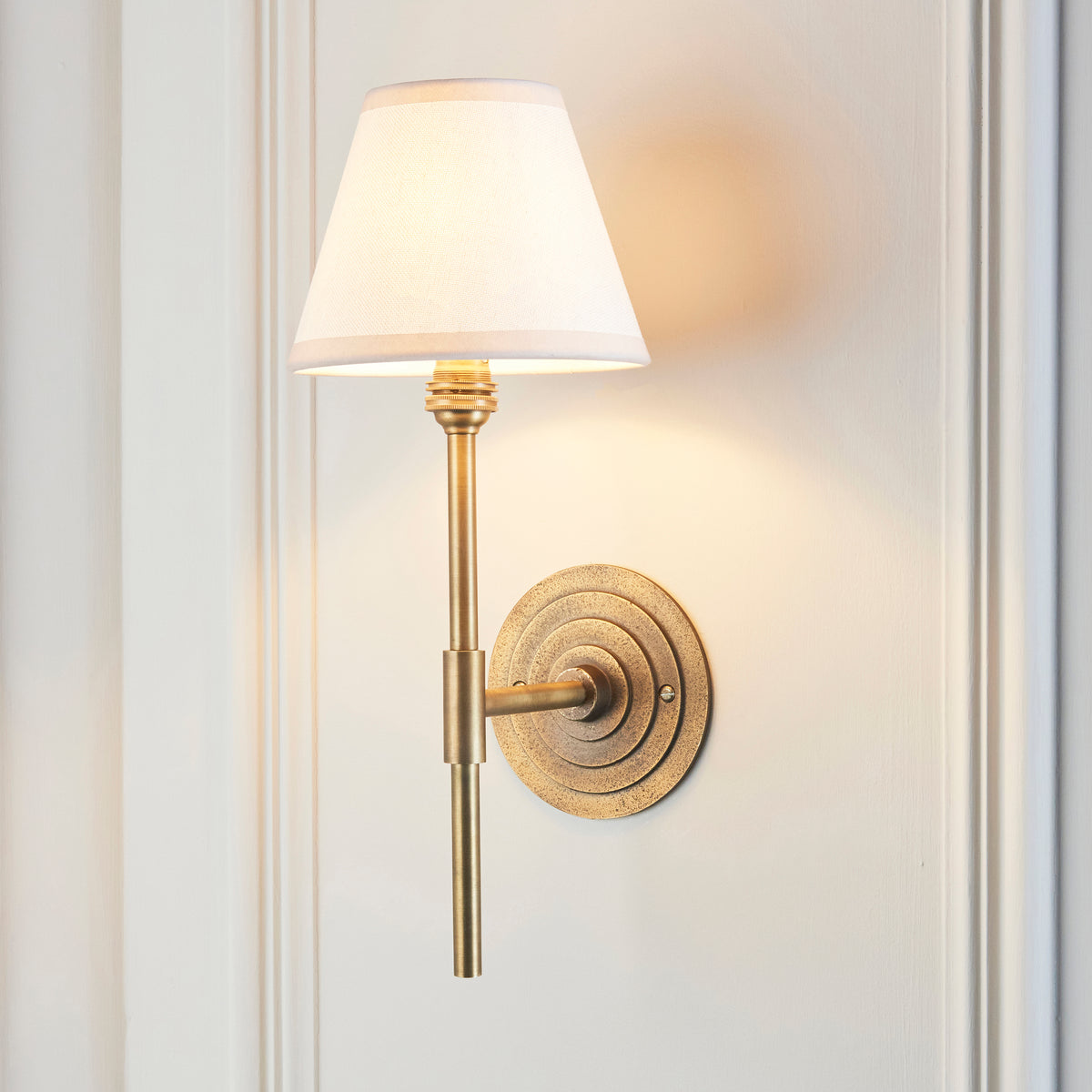 Halo antique brass wall light lit on a white panelled wall, cream shade casting a warm glow