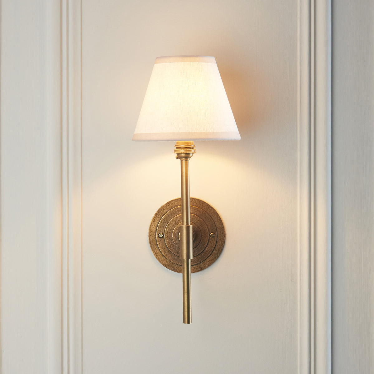 Antique brass Halo wall light illuminated against pale panelling, showing stem and circular backplate