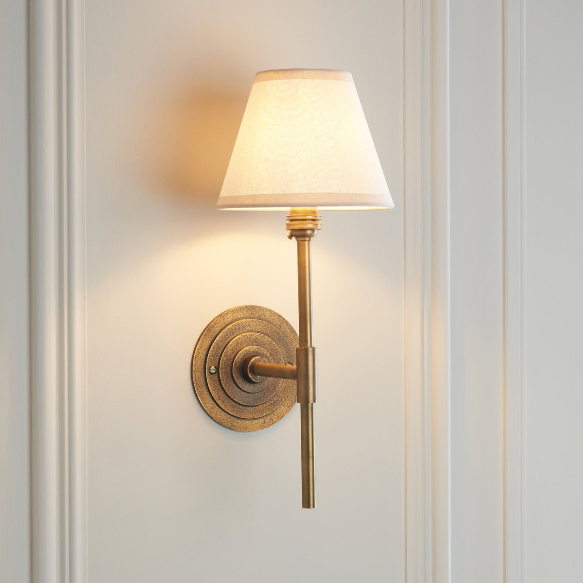 Lit Halo wall light in antique brass with vintage white tapered shade on a cream panelled wall