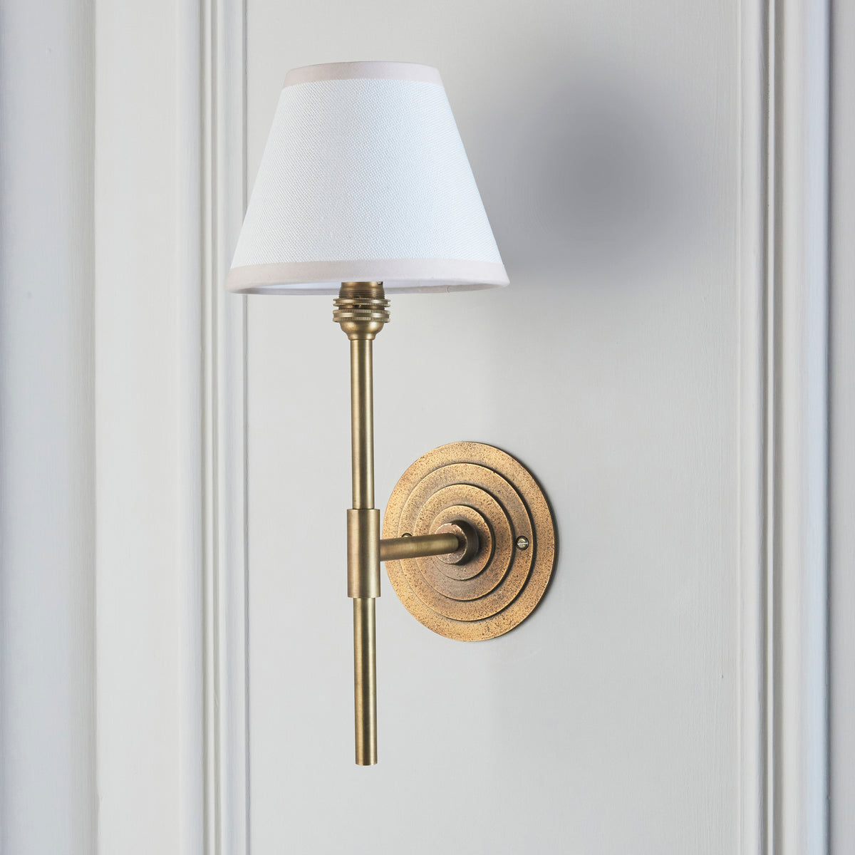 Halo brass wall light switched on, angled view showing stepped backplate and white fabric shade