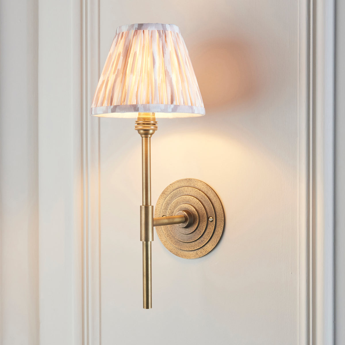Halo Tall brass wall light lit on a white panelled wall, pleated ikat shade glowing softly