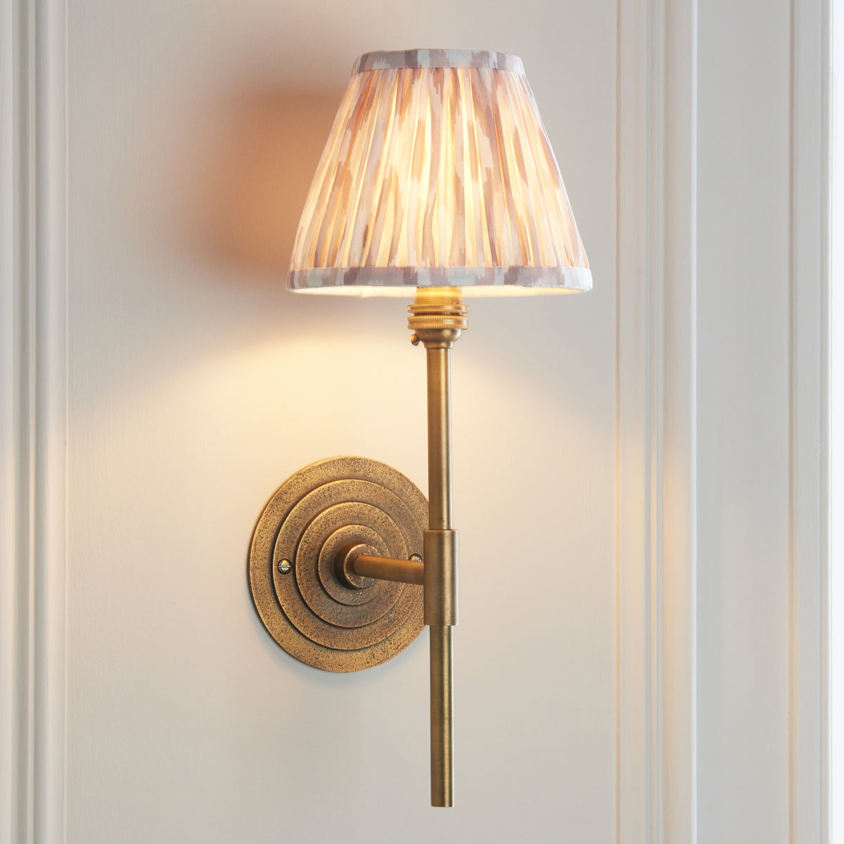Angled view of the lit Halo Tall brass wall light with natural ikat pleated shade