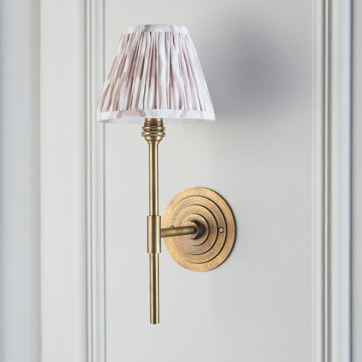 Halo Tall antique brass wall light switched on against a white panelled wall, ikat shade in focus