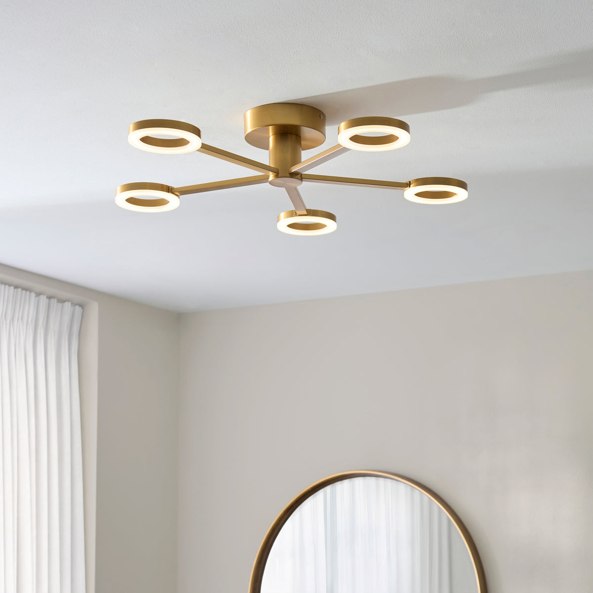 Scout brass LED ring ceiling light lit in a bedroom beside a round mirror and pale curtains