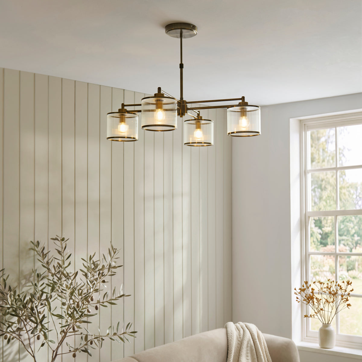 Ridgeton chandelier lit in a living room with green panelled walls, olive tree and cream sofa