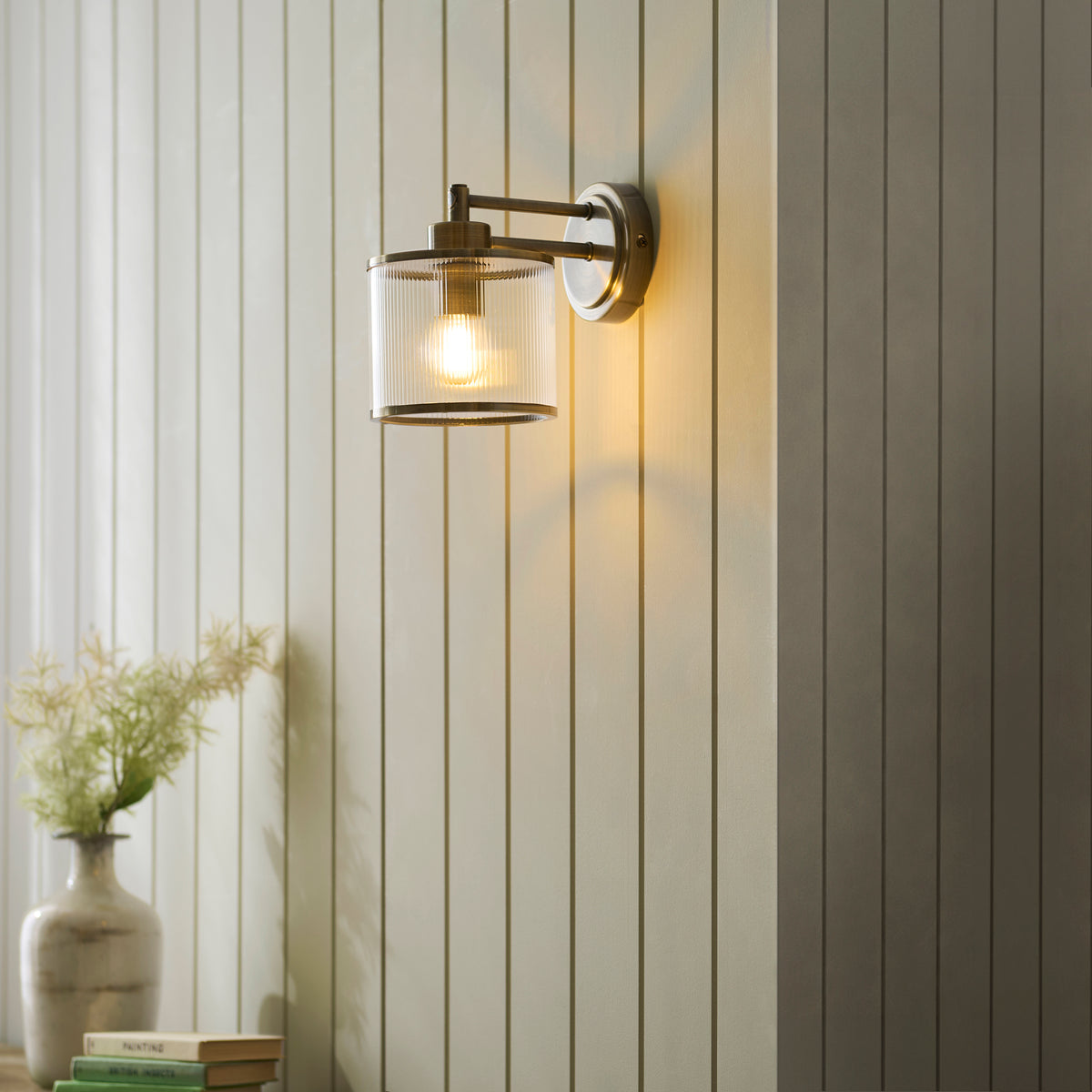 Ridgeton wall light lit on pale panelled wall above books and a stoneware vase of flowers