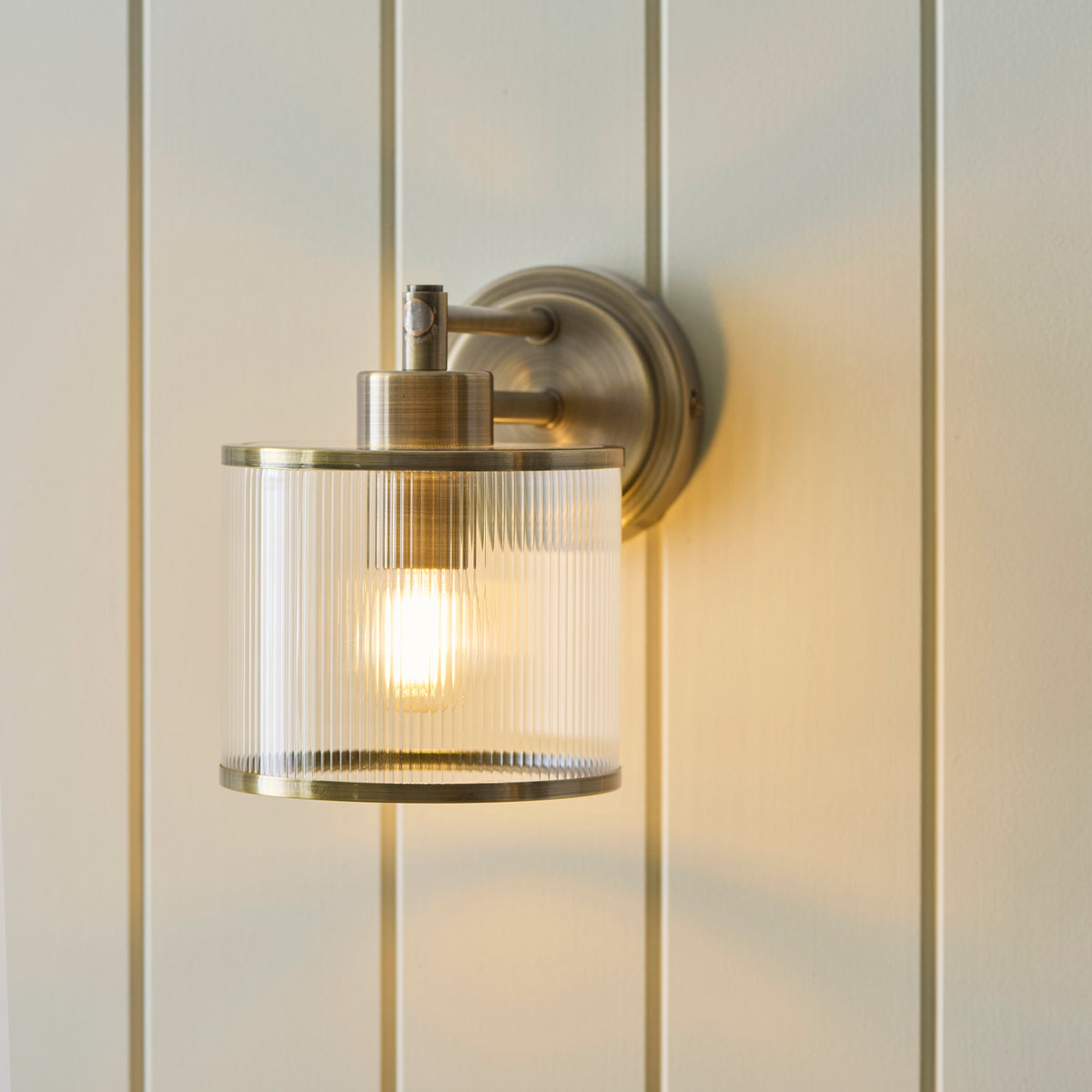 Ridgeton wall light with ribbed glass shade lit on vertical painted panelling