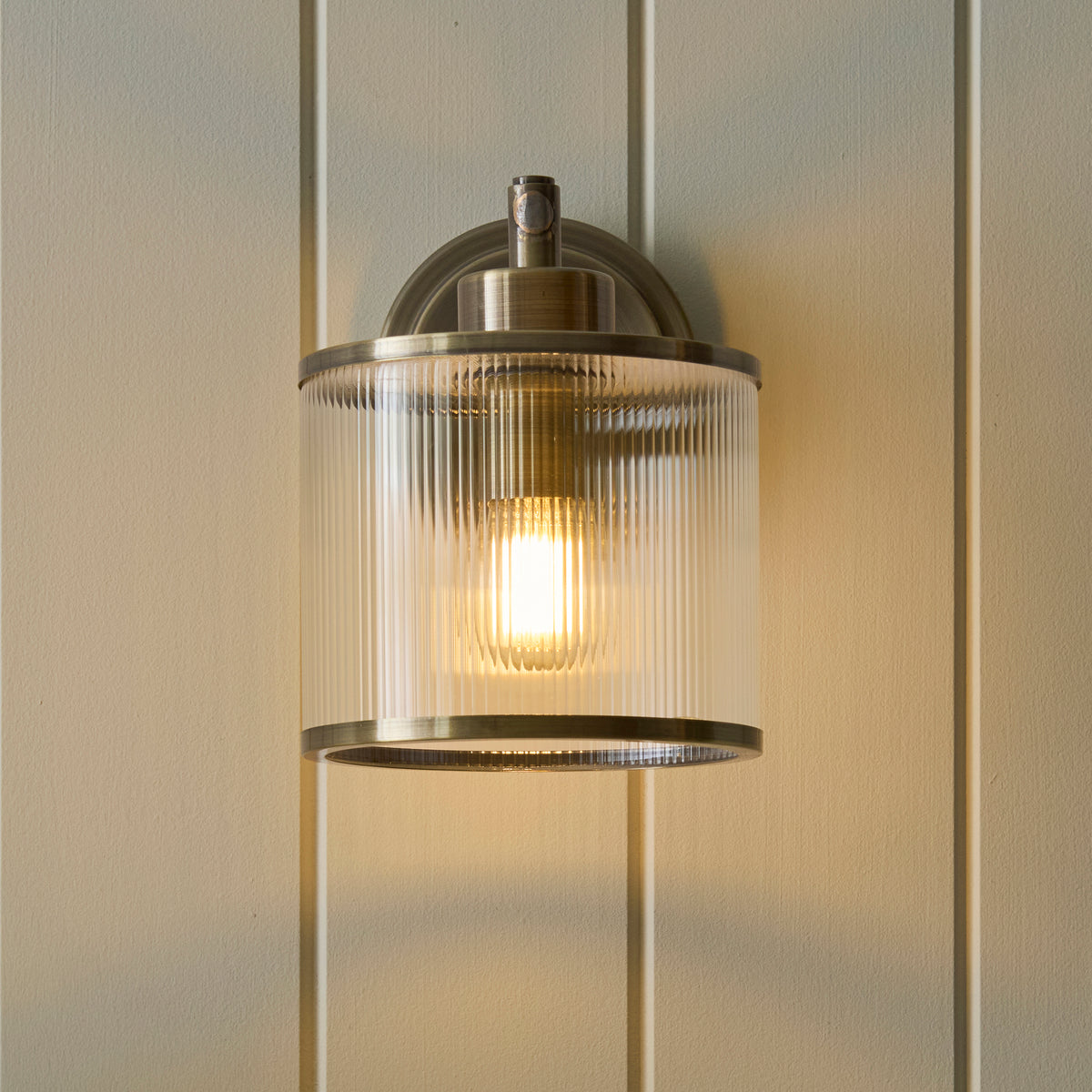 Ridgeton wall light seen from below, its ribbed glass shade glowing warmly on panelled wall
