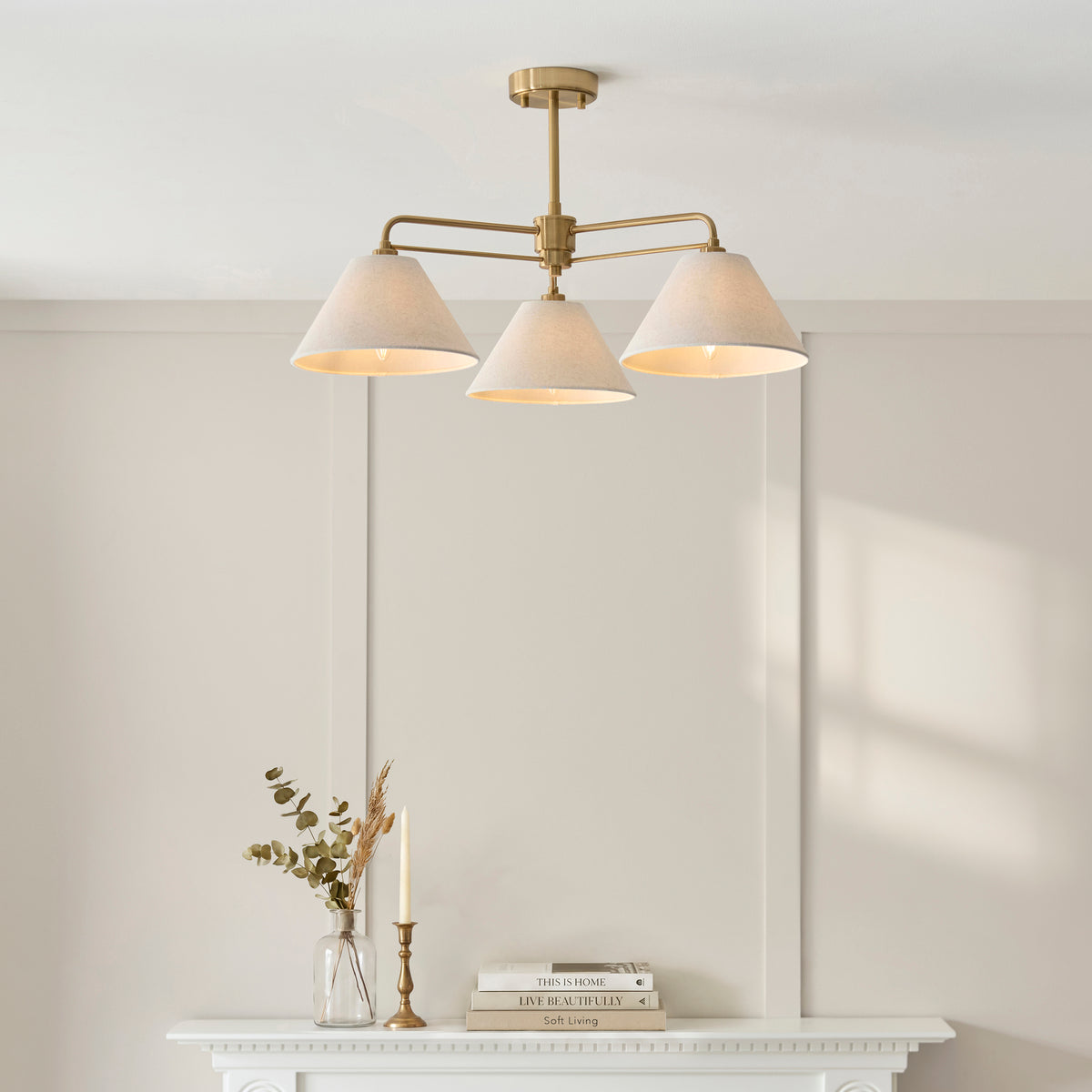 Wallbridge three-light semi flush fitting lit above a white fireplace with eucalyptus and books
