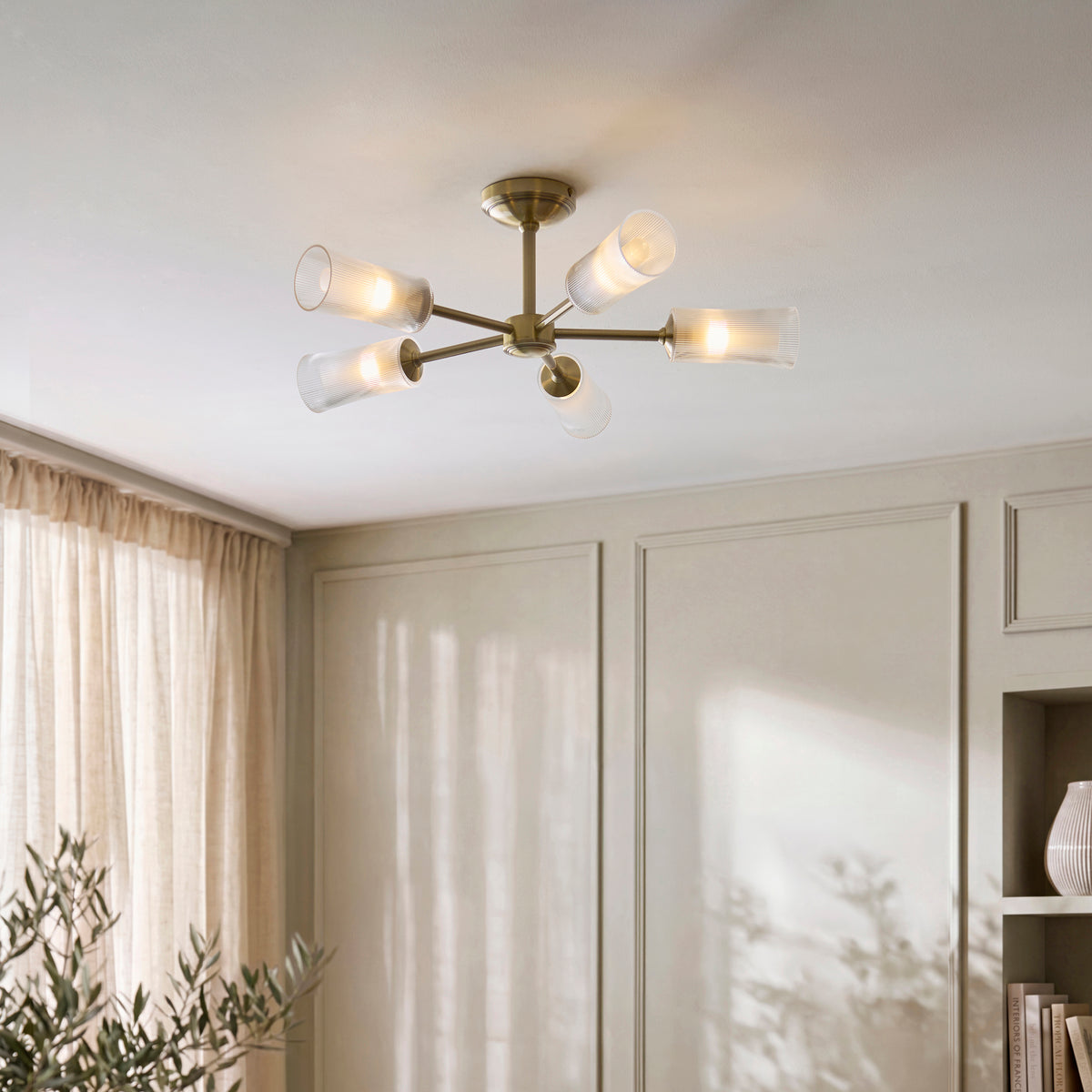 Avelyn brass semi flush light on a ceiling above panelled walls, curtains and shelving
