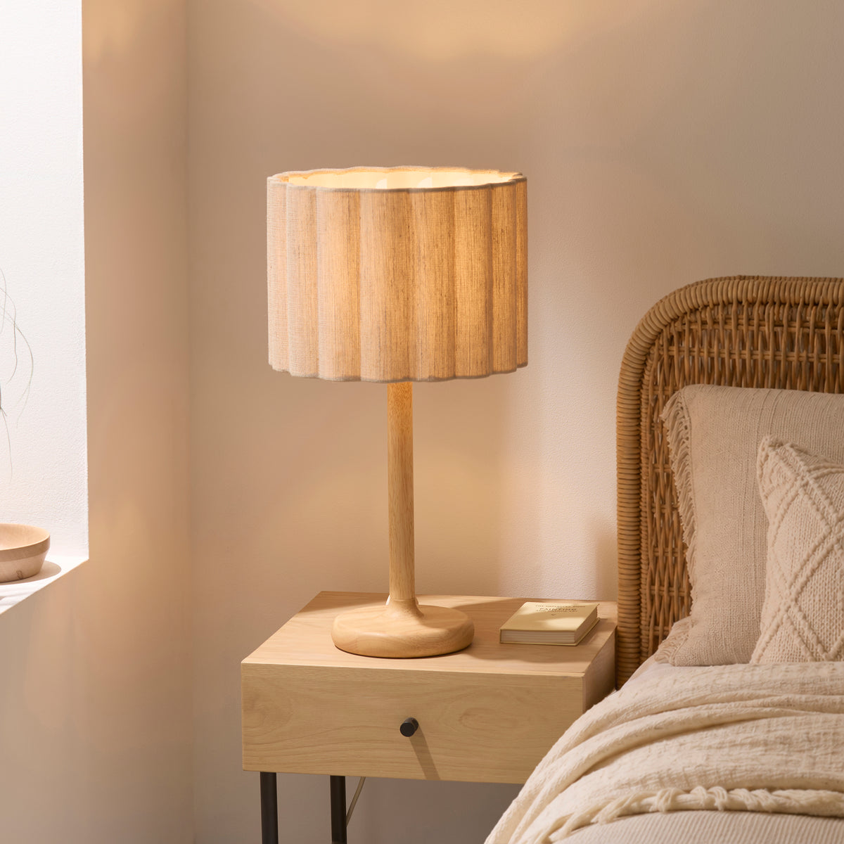 Olson oak table lamp lit on a bedside table beside a rattan headboard and cream knitted cushions