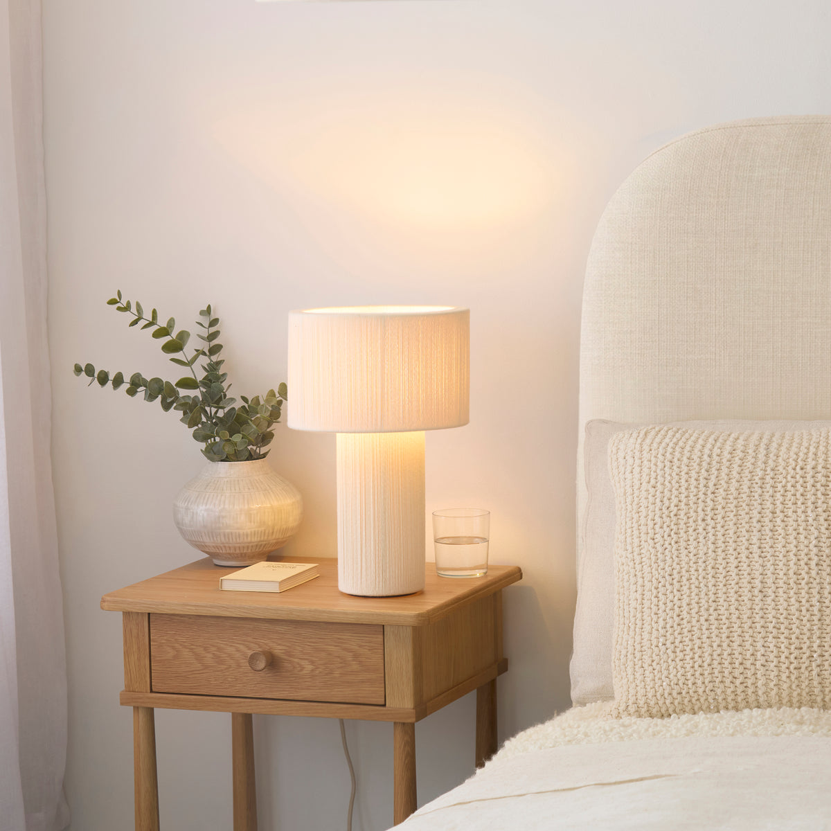 Noah cream cotton table lamp lit on an oak bedside table with eucalyptus in a ribbed vase