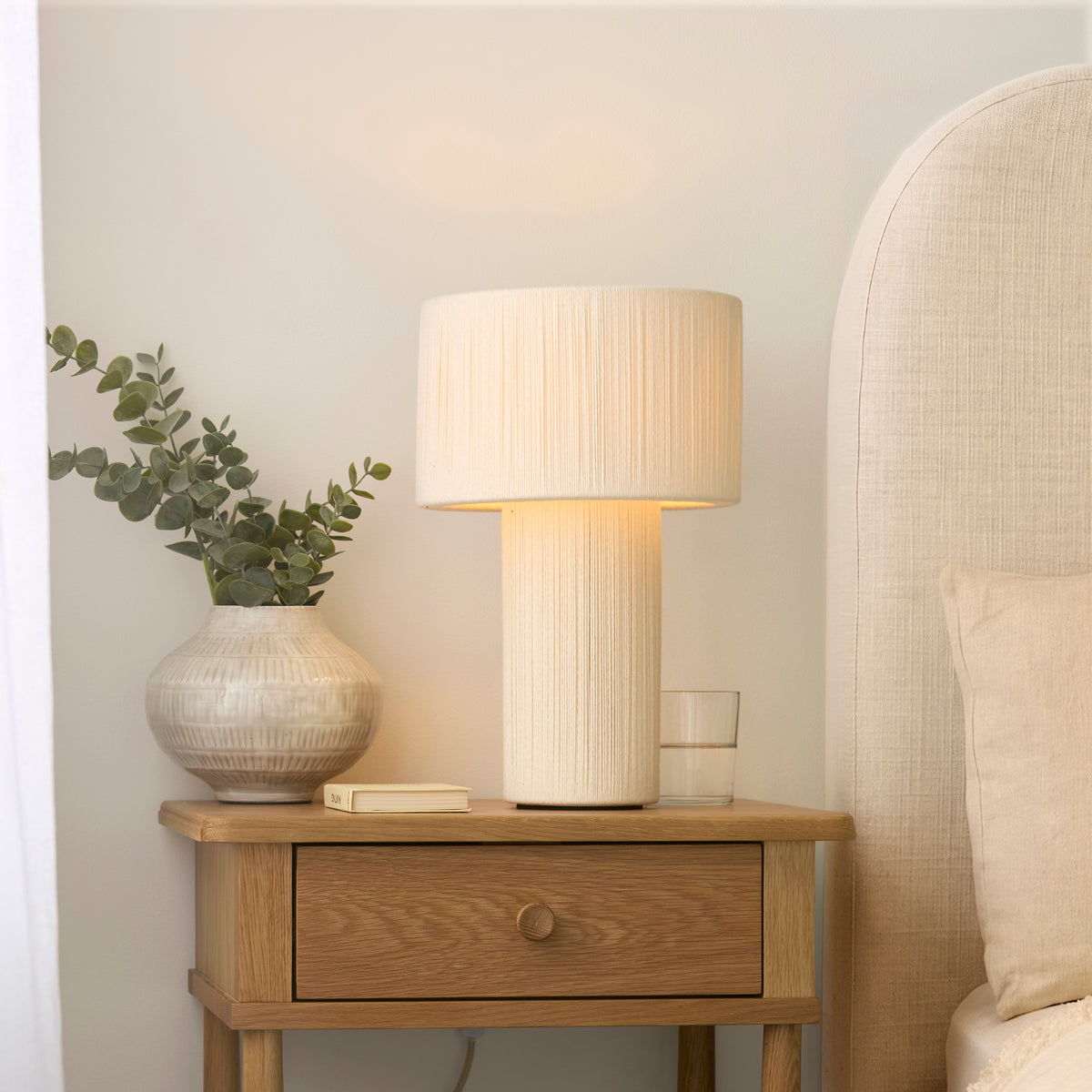 Noah lamp glowing beside a linen headboard, oak drawer unit, book and glass of water