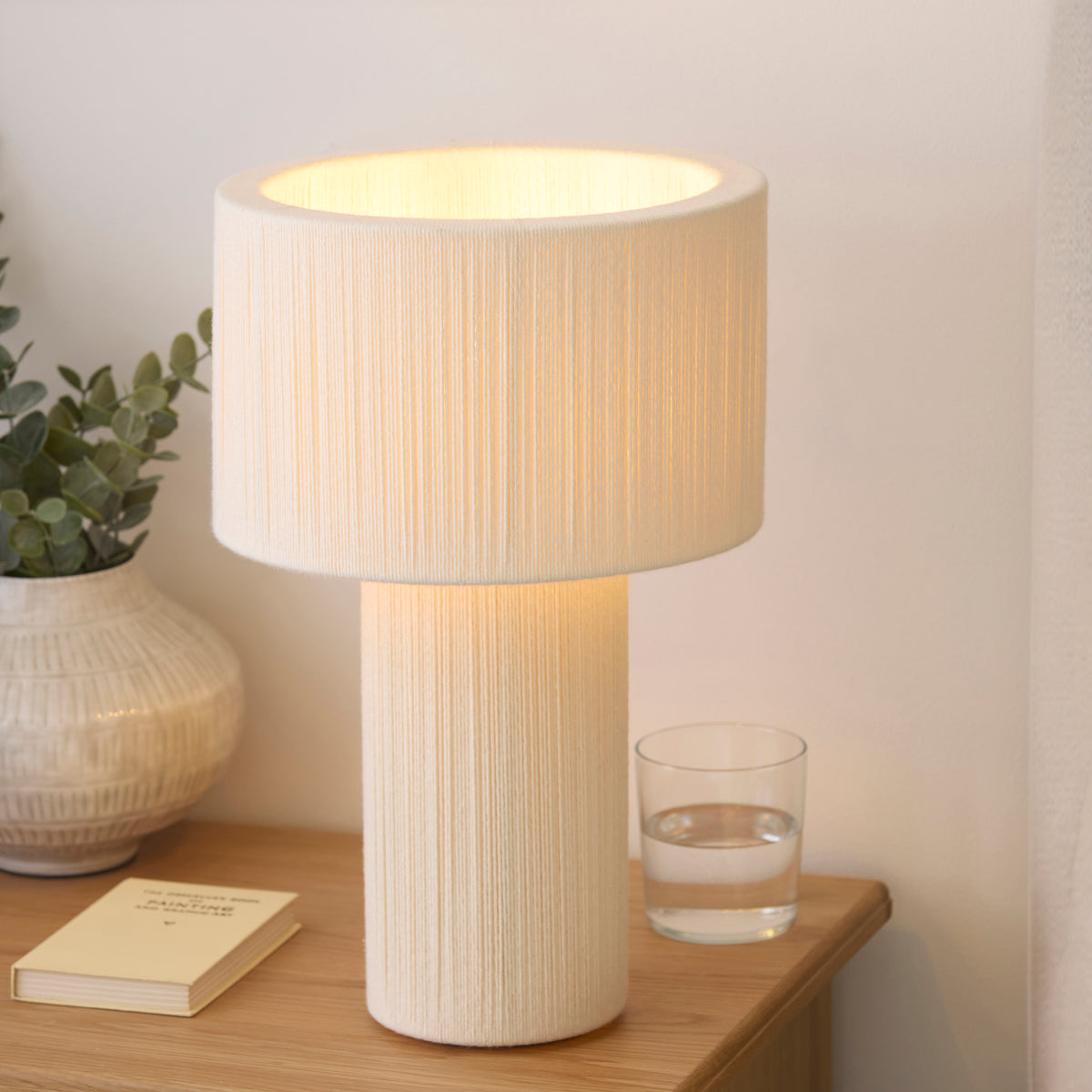 Warmly lit Noah cotton table lamp beside a stoneware vase, book and tumbler on an oak console