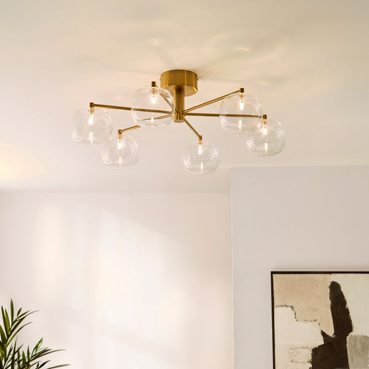 Oslo aged brass six-light semi flush fitting lit on a white ceiling above a framed print and plant