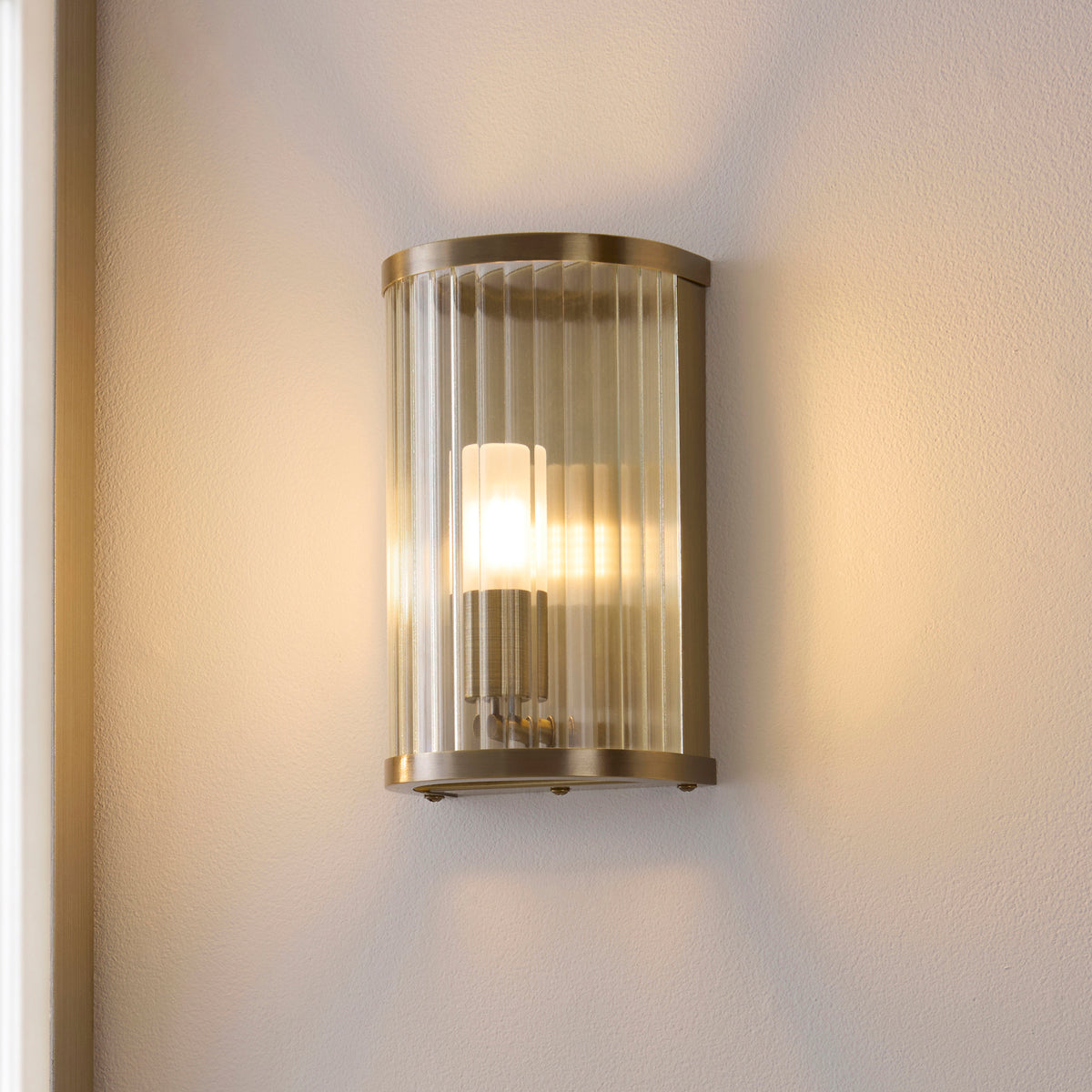 Ridgeton wall light lit on a pale bathroom wall showing its opal inner shade behind ribbed glass