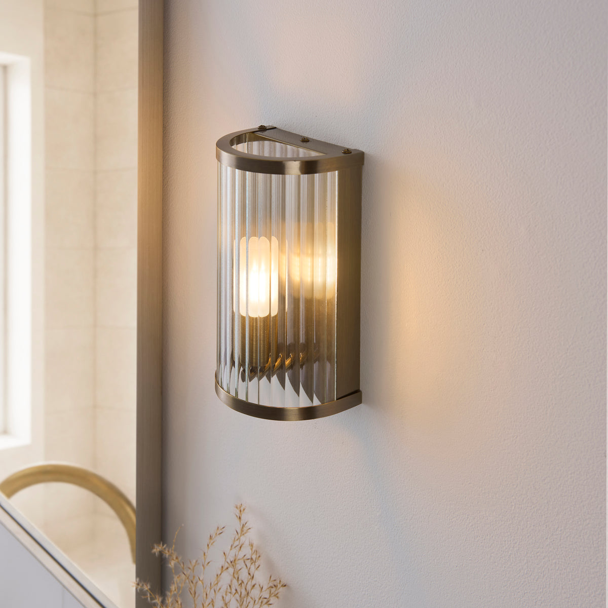 Angled view of the Ridgeton brass wall light beside a bathroom mirror and tiled wall